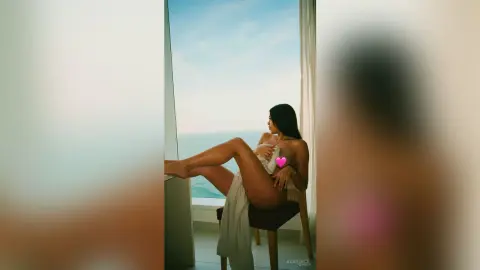Woman in a white summer dress sitting by a balcony overlooking the ocean during a serene sunset.