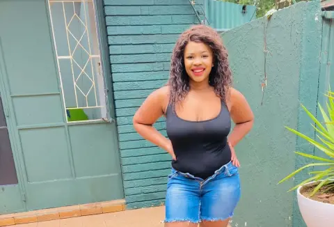 Smiling young woman in a black bodysuit and denim shorts standing outdoors by a green brick wall.