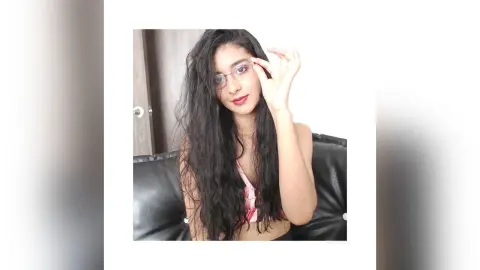 Young woman with long wavy hair and glasses wearing a pink top in a bright indoor setting.