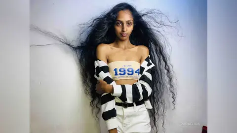 Woman with long wavy hair wearing a black and white striped top and white pants against a plain background.