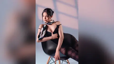 Elegant woman in a black bodysuit and patterned scarf sitting on a chair in soft, cinematic lighting.