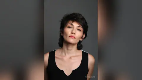 IveteDurand poses looking at the camera wearing a black tank top with her dark hair styled in a short, messy cut against a plain grey background.