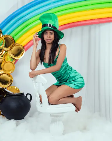 Smiling woman in a green satin dress and leprechaun hat sitting on a white chair in a festive St. Patrick's Day set.