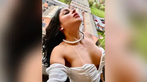 Glamorous woman in a white off-the-shoulder dress and pearl necklace posing on a sunny urban balcony.