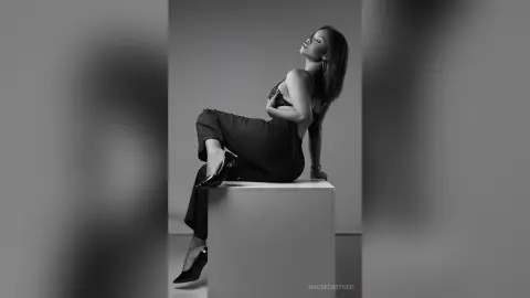 Elegant woman in black trousers and heels posing on a white pedestal in a moody black and white studio portrait.
