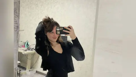 AmberLune poses for a mirror selfie wearing a black tank top and a black zip-up hoodie with her dark hair styled up.