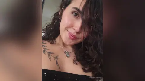 RossiNoleen smiles while wearing a black sparkly top and a silver pendant necklace, showing her shoulder and a leaf tattoo.