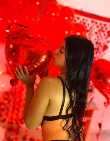 Young woman in black lingerie kissing a large red heart balloon in a festive romantic setting.
