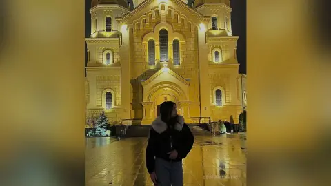 Woman in a black jacket and white faux fur collar standing before a glowing yellow cathedral at night.