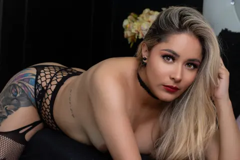 PamelaLeah poses naked with blonde hair, wearing a black choker and black fishnet bottoms against a dark background.