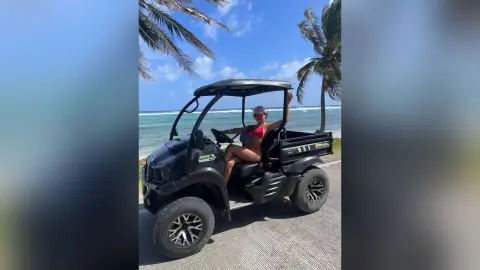 Smiling woman in a red bikini relaxing on a black golf cart at a sunny tropical beach resort.
