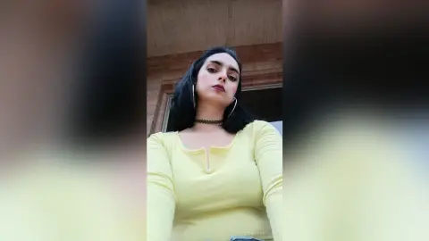 Young woman in a yellow long-sleeve top and black choker posing outdoors with a confident expression.