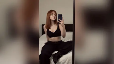 Redheaded woman in a black crop top and baggy trousers taking a moody mirror selfie in a bedroom.