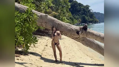 Fit woman in a black bikini standing on a sunny tropical beach next to a large fallen tree trunk.