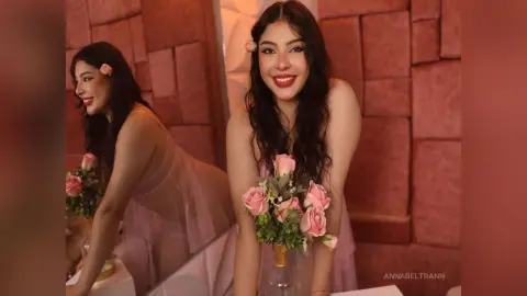 Smiling young woman in a pink tulle dress holding pink roses in a warm, romantic indoor setting.