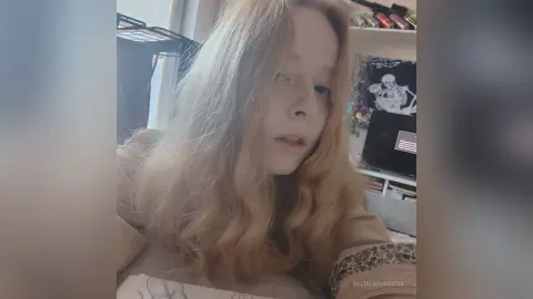Blonde woman in a cozy beige sweater looking thoughtfully at the camera in a bright indoor setting.