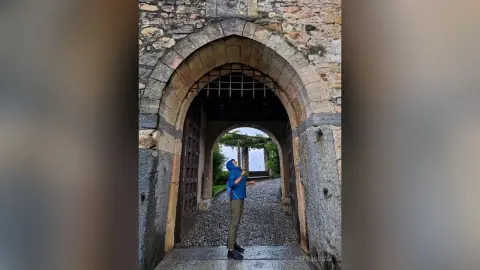 Man in blue hoodie and olive pants standing under a historic stone archway in a peaceful courtyard.