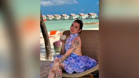 Smiling woman in a blue floral sundress enjoying coffee on a tropical beach resort patio.