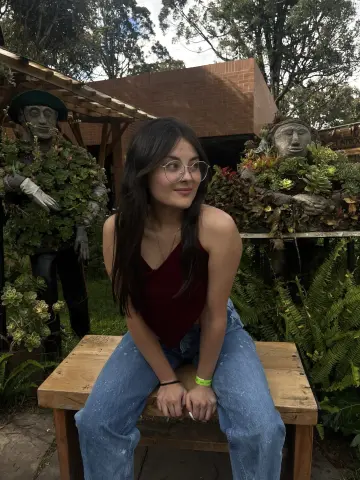 Young woman in a burgundy tank top and jeans sitting on a wooden bench near mossy garden sculptures.
