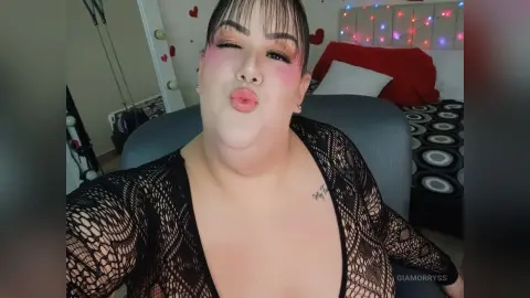 Woman in black lace outfit making a playful kissy face in a cozy bedroom setting.