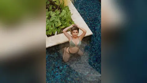 Young woman in a green bikini relaxing in a luxury tiled swimming pool surrounded by lush tropical plants.