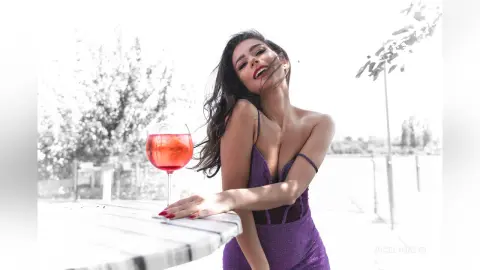 Smiling woman in a purple cocktail dress holding a drink at an outdoor summer garden party.