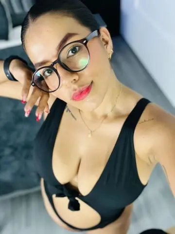 Smiling woman in black swimsuit and glasses posing for a selfie in a bright indoor setting.