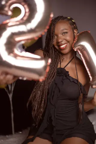 Smiling Black woman in a black ruffle dress celebrating with rose gold number balloons.