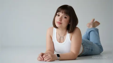 Young woman with short brown hair and chest tattoo wearing a white tank top and jeans, relaxing on a white floor.