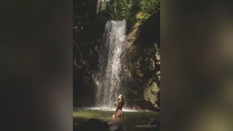 LaurentBelluci stands naked in a shallow pool or stream beneath a tall waterfall in a lush, green forest setting.