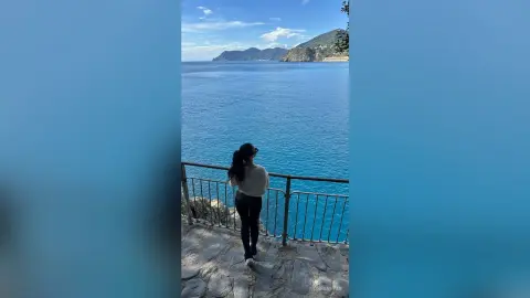 Woman in white top and black pants overlooking the vast blue ocean from a scenic coastal balcony.
