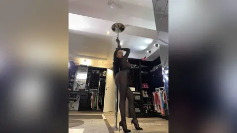 NicoleBella stands upright against a silver pole, wearing a black bodysuit and black high heels.