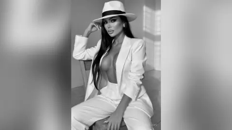 Elegant woman in a white suit and fedora hat posing in a bright, minimalist indoor setting.