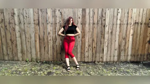 Red-haired woman in a black crop top and red trousers leaning against a rustic wooden fence in soft daylight.