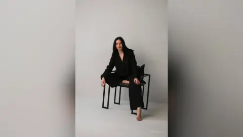 Elegant woman in a black blazer and trousers sitting on a chair against a minimalist white studio background.