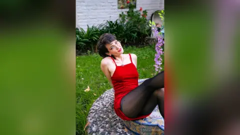 IveteDurand wearing a red dress and black tights, sitting on a stone bench in a garden setting.