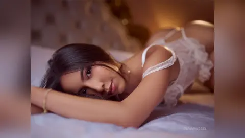 IrinaRussel lies on a bed wearing a white lace teddy, looking at the camera with her dark hair falling over her face.