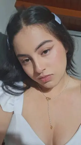 Young woman with dark hair and pearl necklace wearing a white summer top in soft indoor lighting.