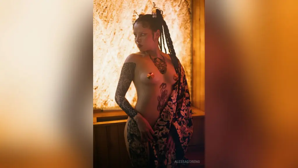 AlessaGoreng — Tattooed woman in traditional patterned wrap and gold headpiece posing in warm, moody lighting.