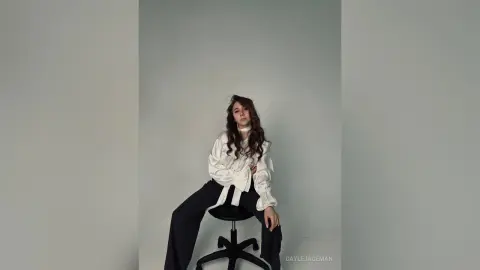 Elegant woman in a white silk blouse and black trousers sitting against a minimalist grey studio background.