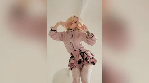 LynGoodhart poses in a pink ruffled dress and white lace stockings, making peace signs with her hands.