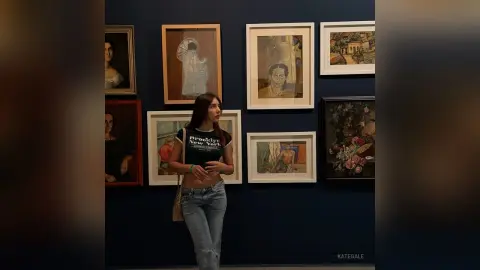 KateGale stands in front of a wall of framed art, wearing a black t-shirt and light blue jeans with her long dark hair down.