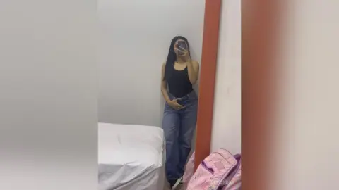Young woman in a black tank top and blue jeans taking a mirror selfie in a minimalist bedroom.