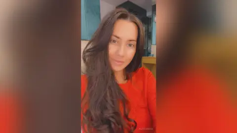 Smiling woman with long wavy brown hair wearing a bright red shirt in a cozy indoor setting.