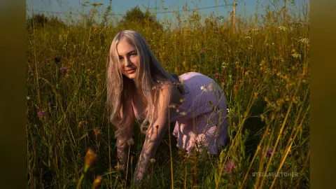 StellaFletcher poses in a grassy field wearing a light pink lace dress with her long blonde hair falling over her shoulders.