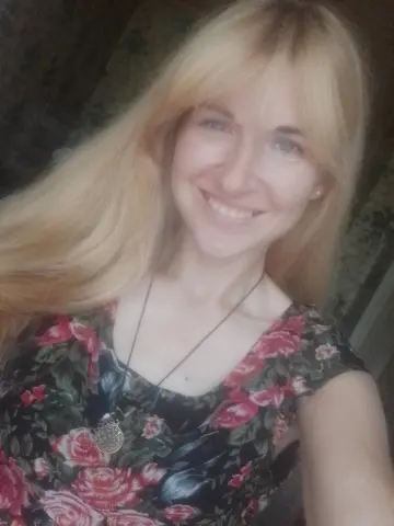 Smiling blonde woman in a floral print blouse and silver necklace posing outdoors in soft natural light.