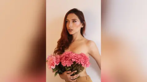 LaurentBelluci holds a large bouquet of pink flowers against her bare chest, looking off to the side with long wavy hair.