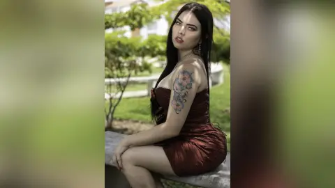 Elegant woman in a dark red satin dress with floral tattoos sitting outdoors in a serene garden setting.