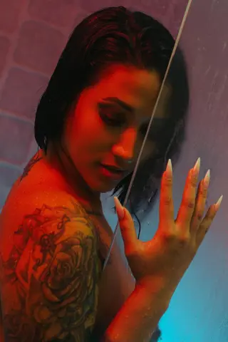 Tattooed woman with wet hair posing against a blue and red neon light background in a moody setting.
