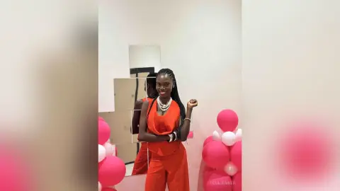 Smiling Black woman in a vibrant red jumpsuit posing in a bright room with pink balloons.